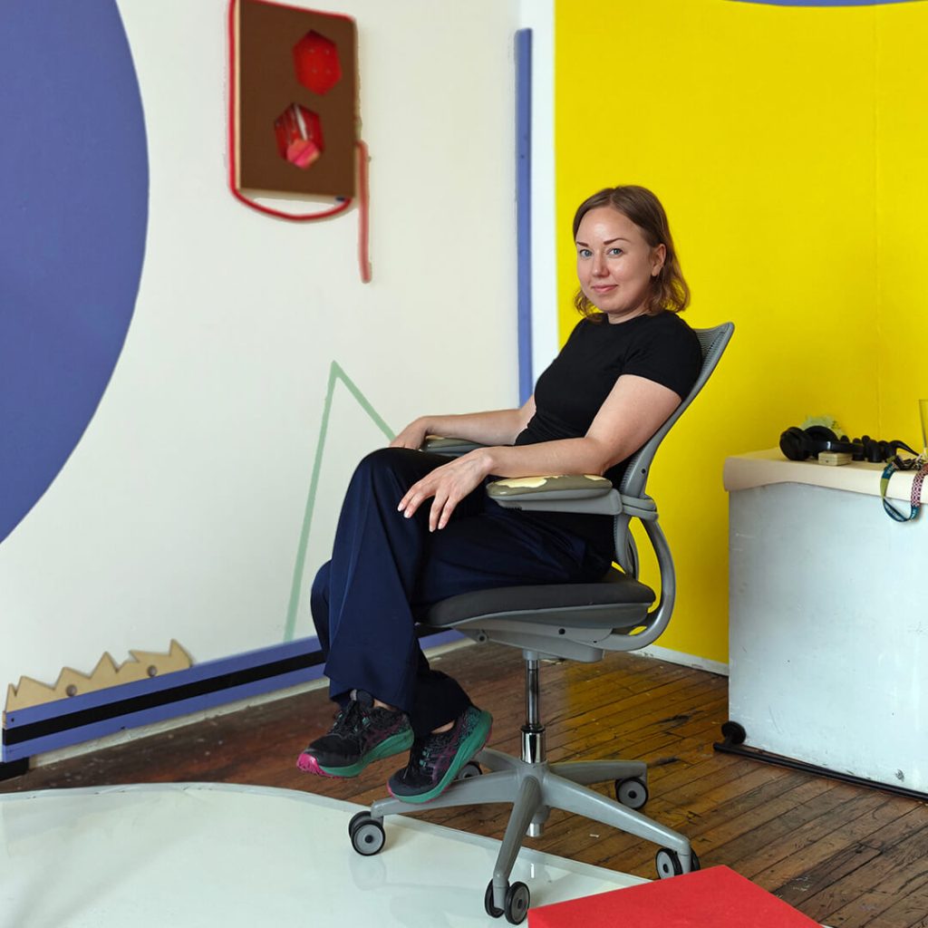 Nainen istuu toimistotuolissa, ympärillä on huoneneen nurkka, jonka seinissä on keltaista sekä muun muassa violetti geometrinen kuvio.