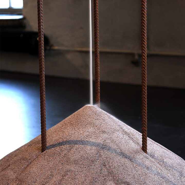 Close-up in the gallery, sand flows in a thin stream from above, gathering in a pile on the floor.