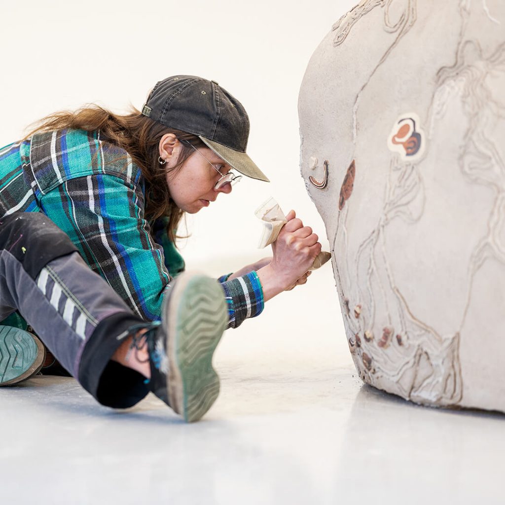 Keskittynyt henkilö istuu maassa ja käsittelee möykkymäisen objektin pintaa.