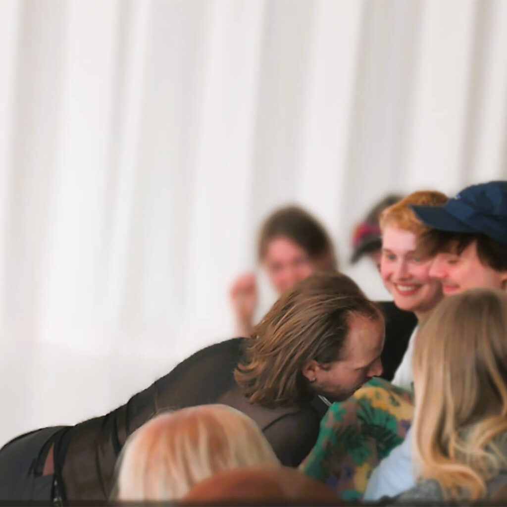 Tummiin vaatteisiin pukeutunut esiintyjä on osittain makuulla, nojautuu kohti ihmisiä ja on vuorovaikutuksessa tyytyväisen näköisen yleisönsä kanssa.