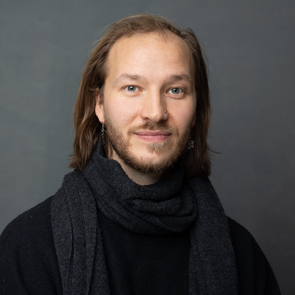 Mustaan paitaan ja harmaaseen huiviin pukeutunut henkilö, jolla on partaa ja puolipitkät hiukset kuvattuna harmaata taustaa vasten.