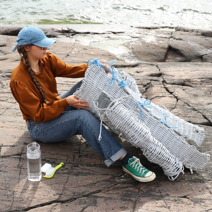 Lippalakkiin, murretun oranssiin paitaan ja farkkuihin pukeutunut henkilö istuu rantakallioilla ja pitelee käsissään valkoisesta rottinkimaisesta materiaalista tehtyä teoksen osaa.