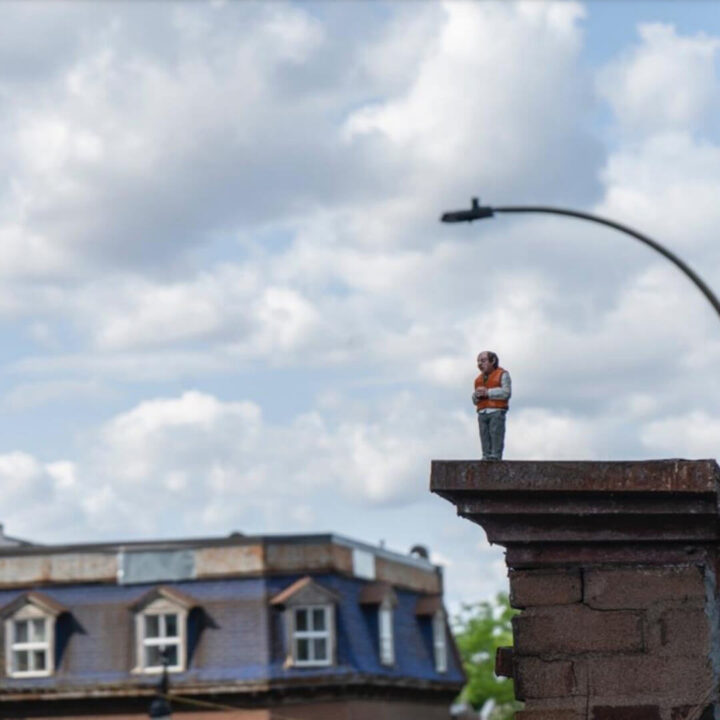 A small human figure dressed in a red vest and gray pants stands on top of a tall roof structure. Clouds and blue sky and the attic of another house in the background.