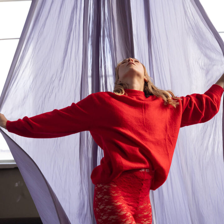 Punaisiin vaatteisiin pukeutunut vaaleahiuksinen nainen seisoo ylhäältä laskeutuvan vaaleanvioletin kankaan edessä ja levittää sitä käsillään.