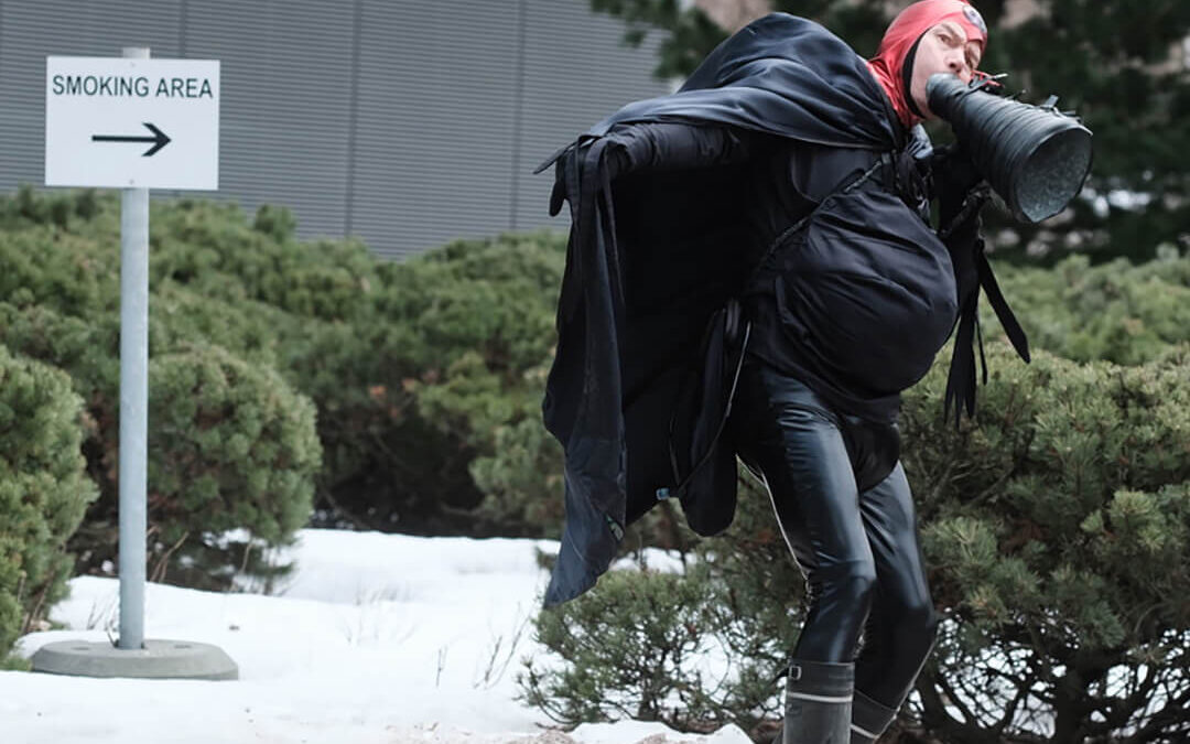 Mustiin vaatteisiin ja viittaan pukeutunut Tulipää-hahmo pitelee mustaa megafoni-esinettä. Taustalla pensasaita ja "Smoking area" -kyltti.