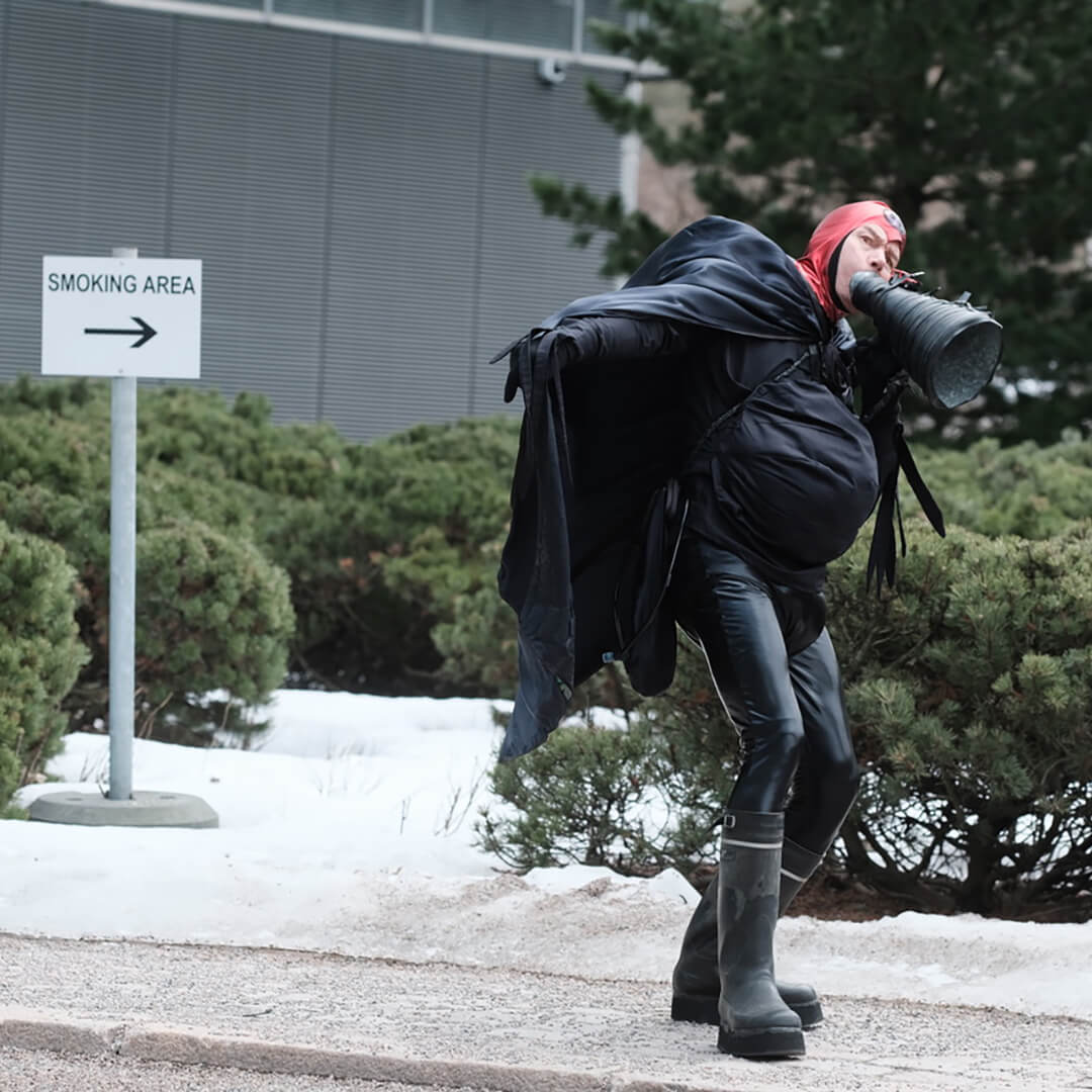 Mustiin vaatteisiin ja viittaan pukeutunut Tulipää-hahmo pitelee mustaa megafoni-esinettä. Taustalla pensasaita ja "Smoking area" -kyltti.