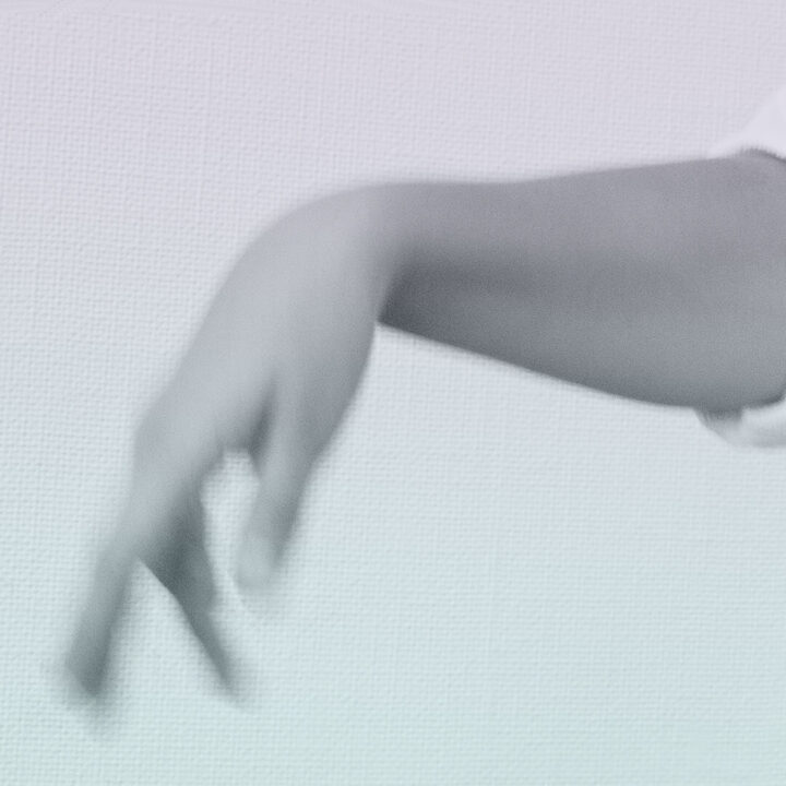 Valkoiseen paitaan puetun henkilön käsi kuvattuna vaaleaa taustaa vasten, käsi on vaakasuorassa ja sormet roikkuvat rennosti. Kuvassa on mustavalkoisen kuvan tuntu, ja väreinä harmaasävyjen lisäksi muun muassa hentoa turkoosia ja lilaa.