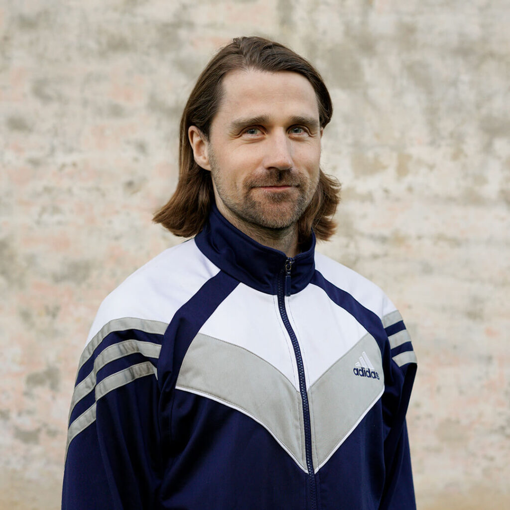 A bearded man wearing a blue and white Adidas tracksuit jacket smiles and looks at the camera. Photographed against a light, slightly uneven background.