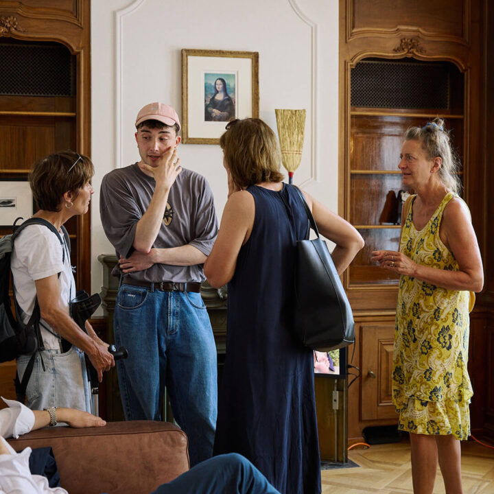A small group of people are gathered to chat in an interior space with wooden cabinets that look dignified and old-fashioned. The person in the middle is wearing a pink cap, a gray shirt, and jeans, and the others are looking at him.
