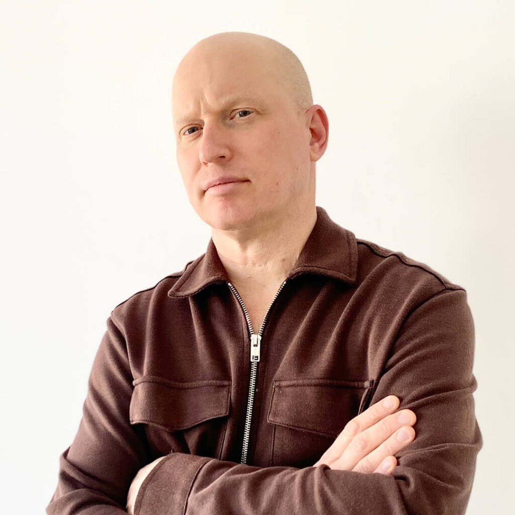 A serious-looking, bald, light-skinned man wearing a brown long-sleeved shirt looks at the camera.