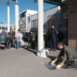 Kaupunkitila, jossa ihmiset katsovat vihrtävään kokohaalariin pukeutunutta miestä, joka on kyykyssä metallilevyn päällä työhanskat kädessään.