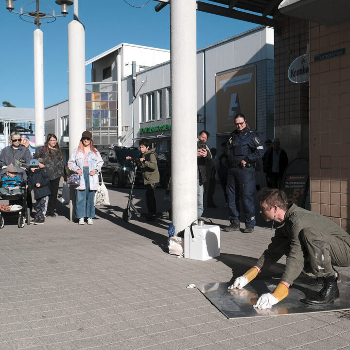Kaupunkitila, jossa ihmiset katsovat vihrtävään kokohaalariin pukeutunutta miestä, joka on kyykyssä metallilevyn päällä työhanskat kädessään.